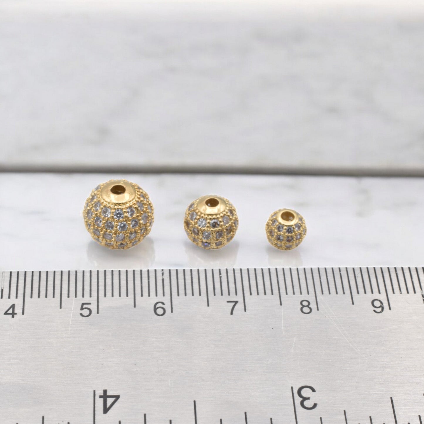 Three gold pave spacer beads with cubic zirconia next to a ruler.