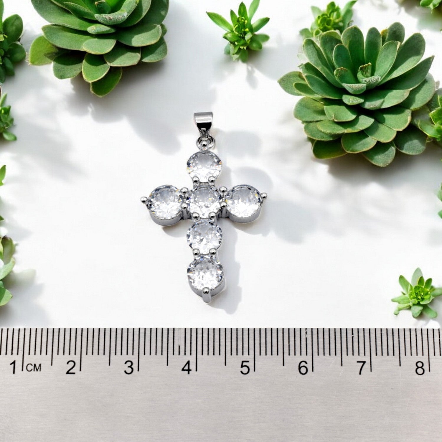 Silver cross pendant with crystal gems next to a ruler and succulents.