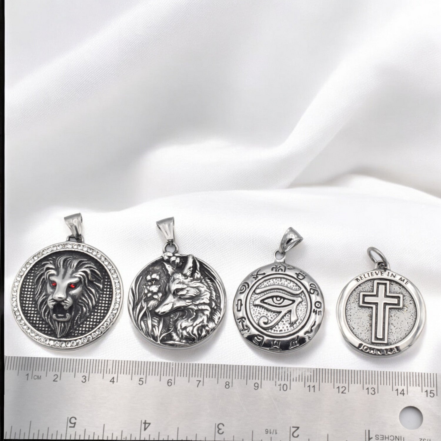 Four silver round pendants with lion, wolf, Eye of Ra, and cross designs displayed on white fabric next to a ruler.